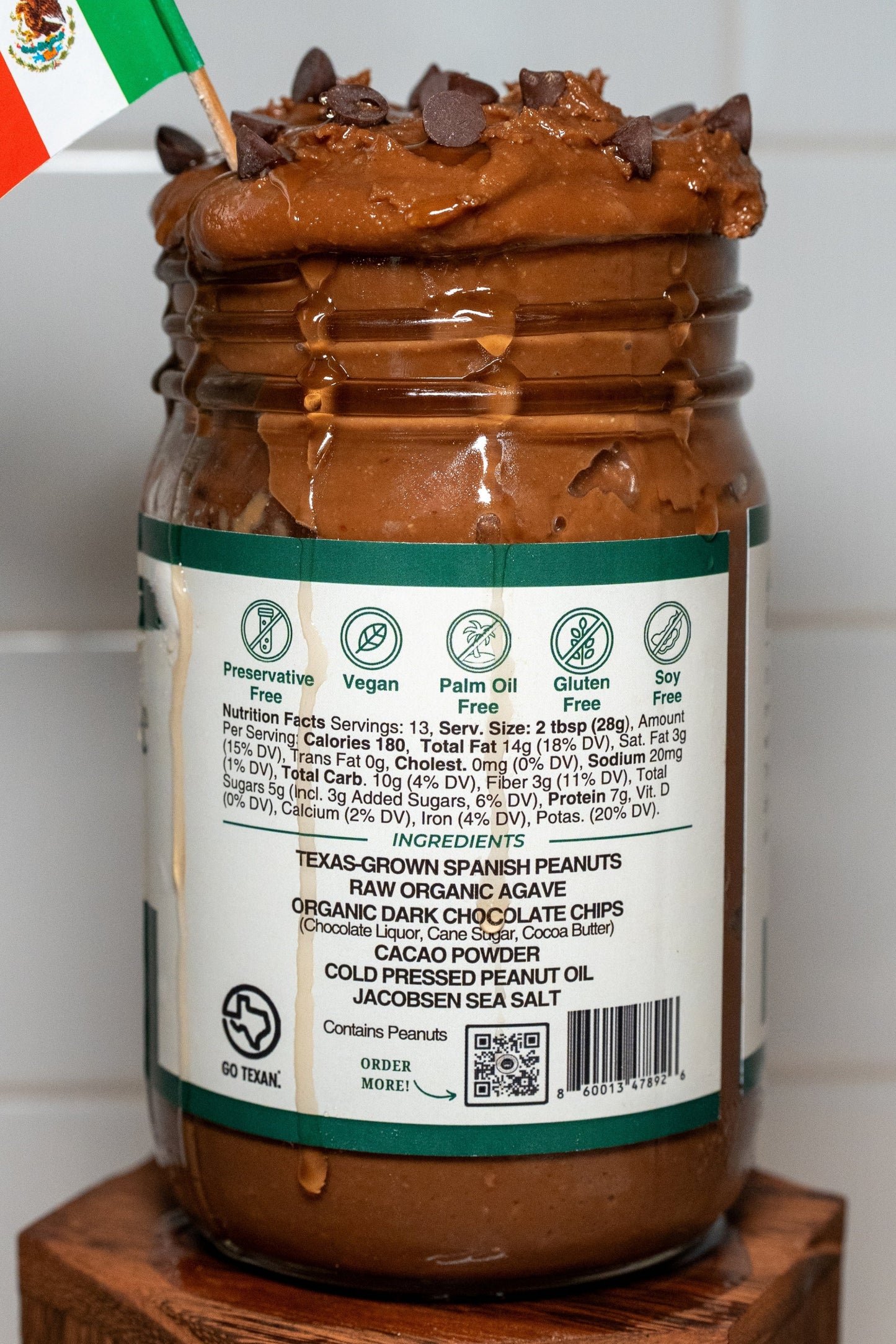 Side photo showing ingredients of a jar of Cacao & Agave Peanut Butter with a Mexican flag on top against a white tiled wall.