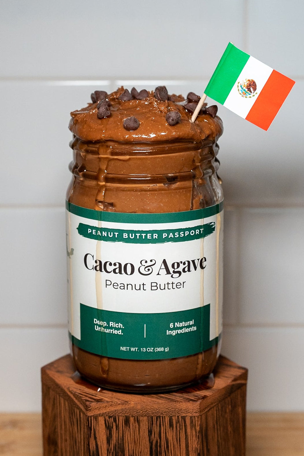 Jar of Cacao & Agave peanut butter on a wooden block with a Mexican flag decoration against a white tiled wall.
