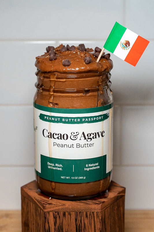 Jar of Cacao & Agave peanut butter on a wooden block with a Mexican flag decoration against a white tiled wall.
