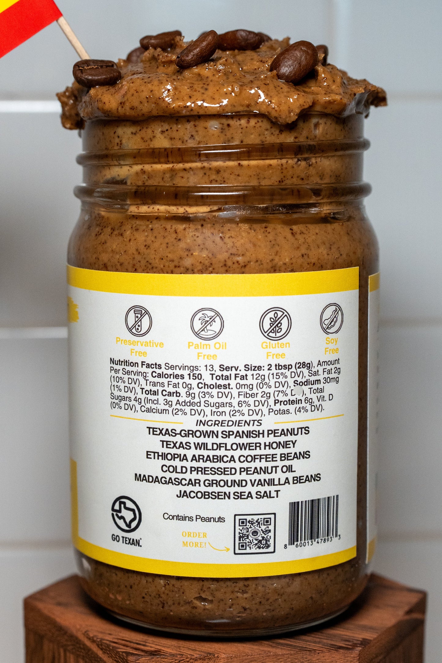 Jar of espresso peanut butter with an ingredient label, placed on a wooden block against a white tiled wall.