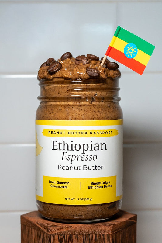 Jar of Ethiopian Espresso Peanut Butter on a wooden stand with a colorful flag on a white tiled wall background.