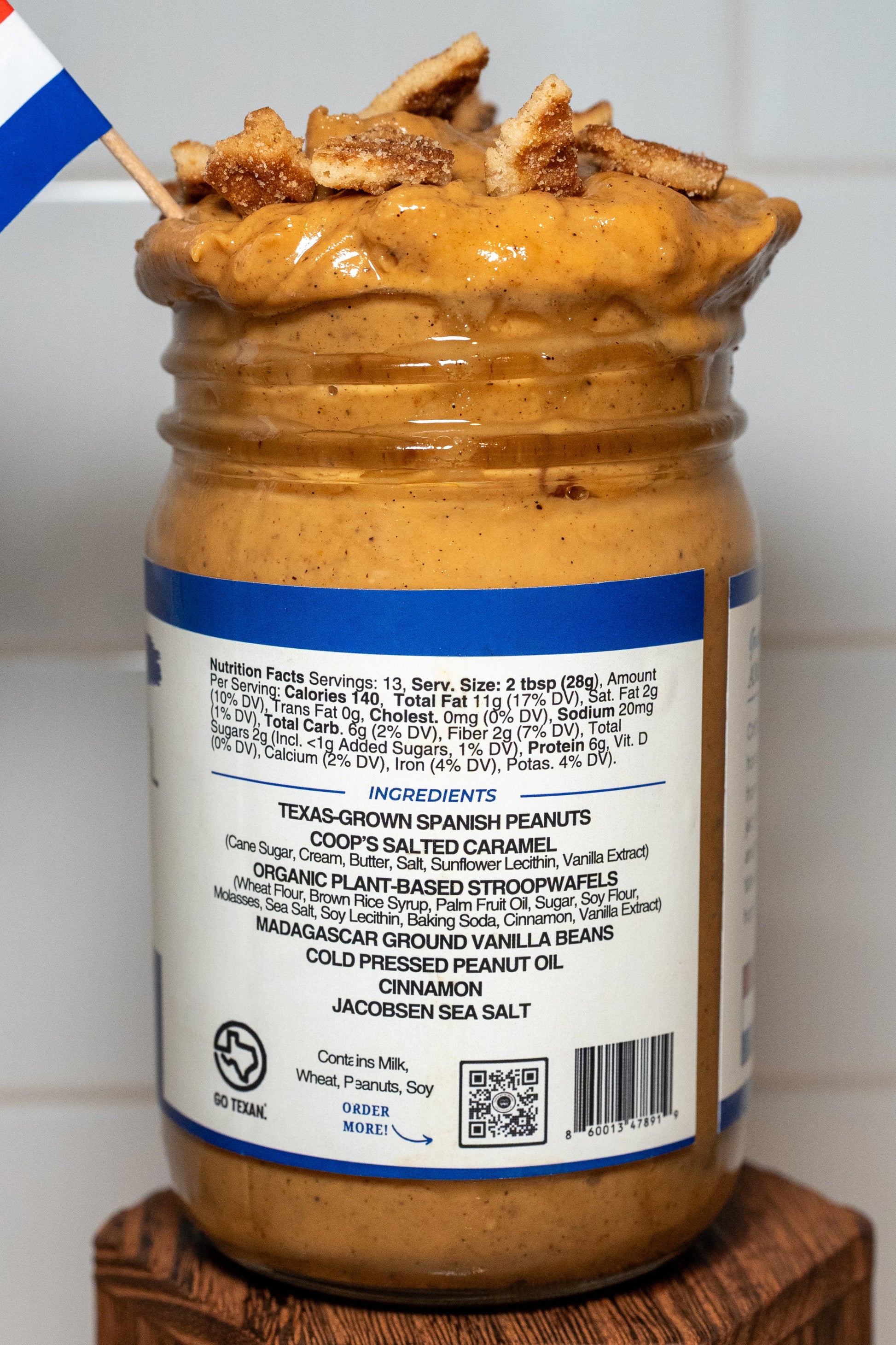 Jar of peanut butter with a label on a wooden block against a tiled wall.