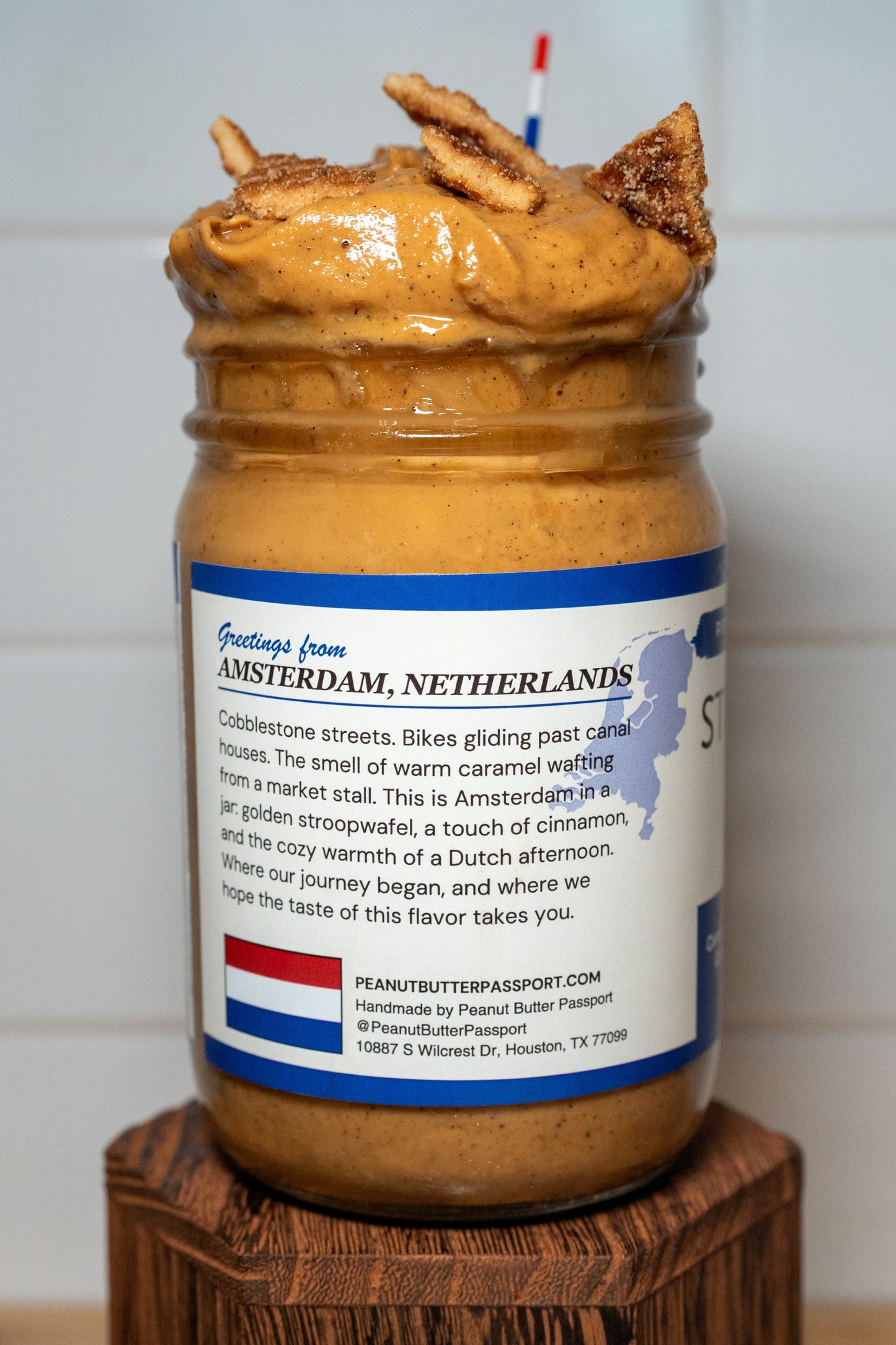 Jar of peanut butter with a label on a wooden block against a white tiled wall.