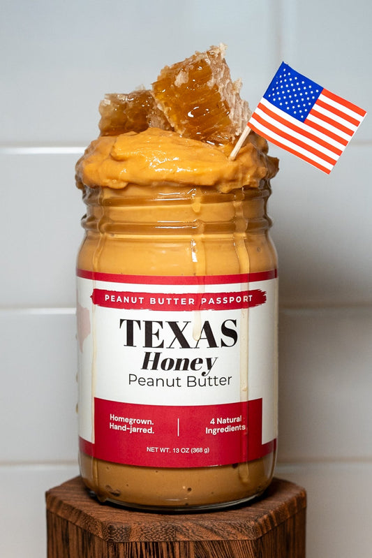 Jar of Texas Honey Peanut Butter on a wooden stand with a small American flag on a white tiled wall background.