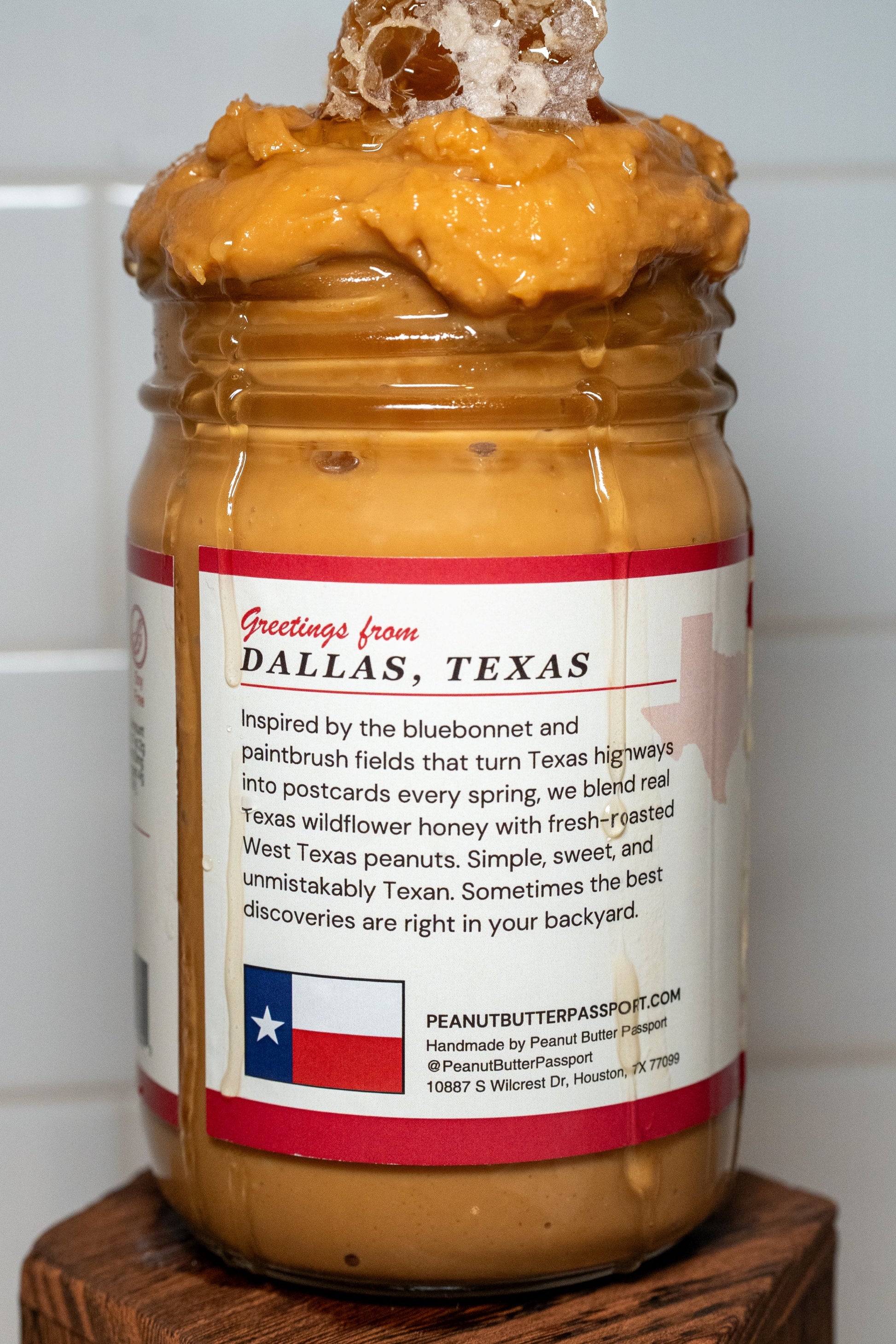 Jar of Texas honey peanut butter with a decorative top on a wooden block against a tiled wall.