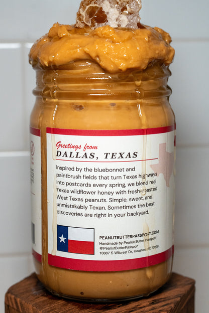 Jar of Texas honey peanut butter with a decorative top on a wooden block against a tiled wall.